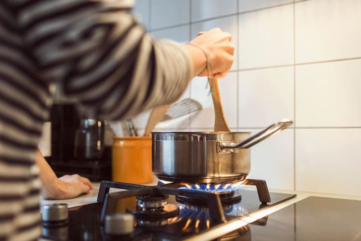 A person wearing a striped shirt stirs food in a pot on a gas stove with a wooden spoon in a tiled kitchen.
